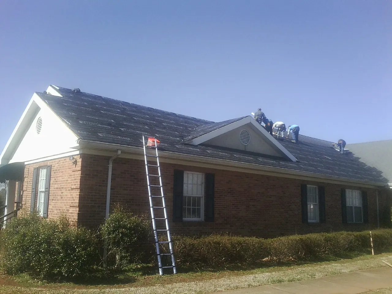 Crew tearing shingles off a brick ranch roof with a ladder set — Emergency Roof Repair work by our crew in Interlochen