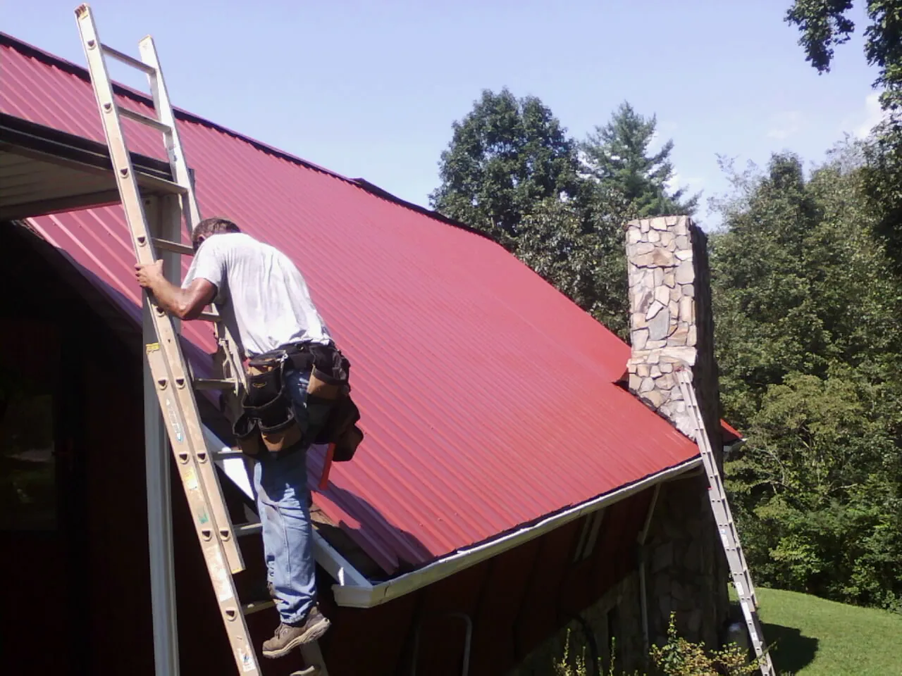 Roofer on a ladder beside a red metal roof and stone chimney — Soffit & Fascia Repair by Roofing Brothers in Interlochen